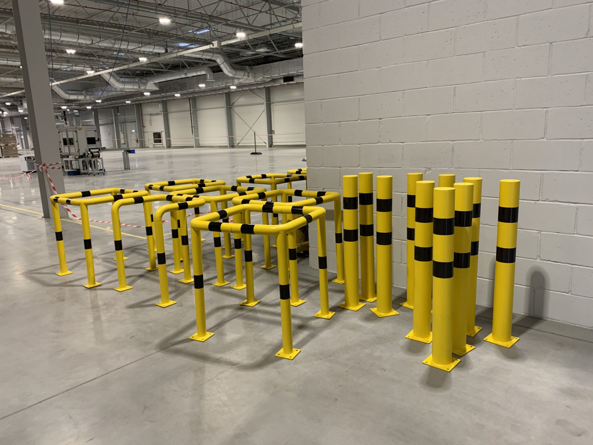 Żółte bariery ochronne z czarnymi pasami w hali magazynowej, widoczne filary konstrukcyjne i instalacje pod sufitem.