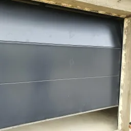 Fragment szarej, segmentowej bramy garażowej w trakcie montażu, widoczne ślady dłoni na panelach, surowy betonowy ościeżnicy z odpryskami.