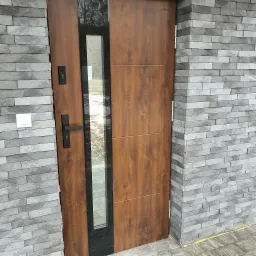 Brązowe drzwi wejściowe z czarnym, pionowym przeszkleniem, zamontowane w elewacji z szarej cegły klinkierowej. Czarny, prostokątny uchwyt i zamek na drzwiach. Widoczny fragment chodnika z kostki...