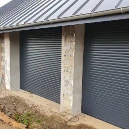 Szare, zamknięte rolety na budynku w trakcie wykończenia, widoczne niedokończone ściany i teren wokół.