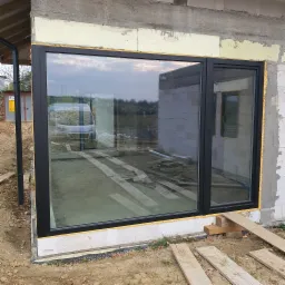 Montaż okna z czarną ramą w surowym stanie budynku, widoczna piana montażowa, odbicie samochodu dostawczego w szybie.