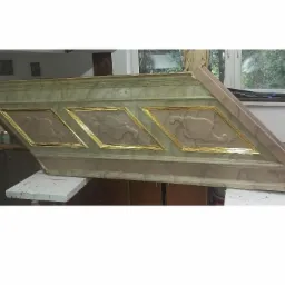 Fragment odrestaurowanej, drewnianej dekoracji architektonicznej z trzema romboidalnymi polami zdobionymi złoceniami, leżący na stole w warsztacie.