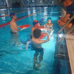 Grupa dzieci i dorosły w basenie podczas zajęć, używają piankowych makaronów i desek do pływania, instruktor siedzi na brzegu basenu.