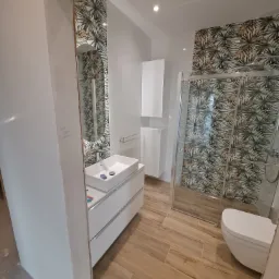 Nowoczesna łazienka z drewnopodobną podłogą, białą zabudową umywalki i kabiną prysznicową z wzorzystą ścianą w motywy roślinne, widok przez surową ościeżnicę drzwi.