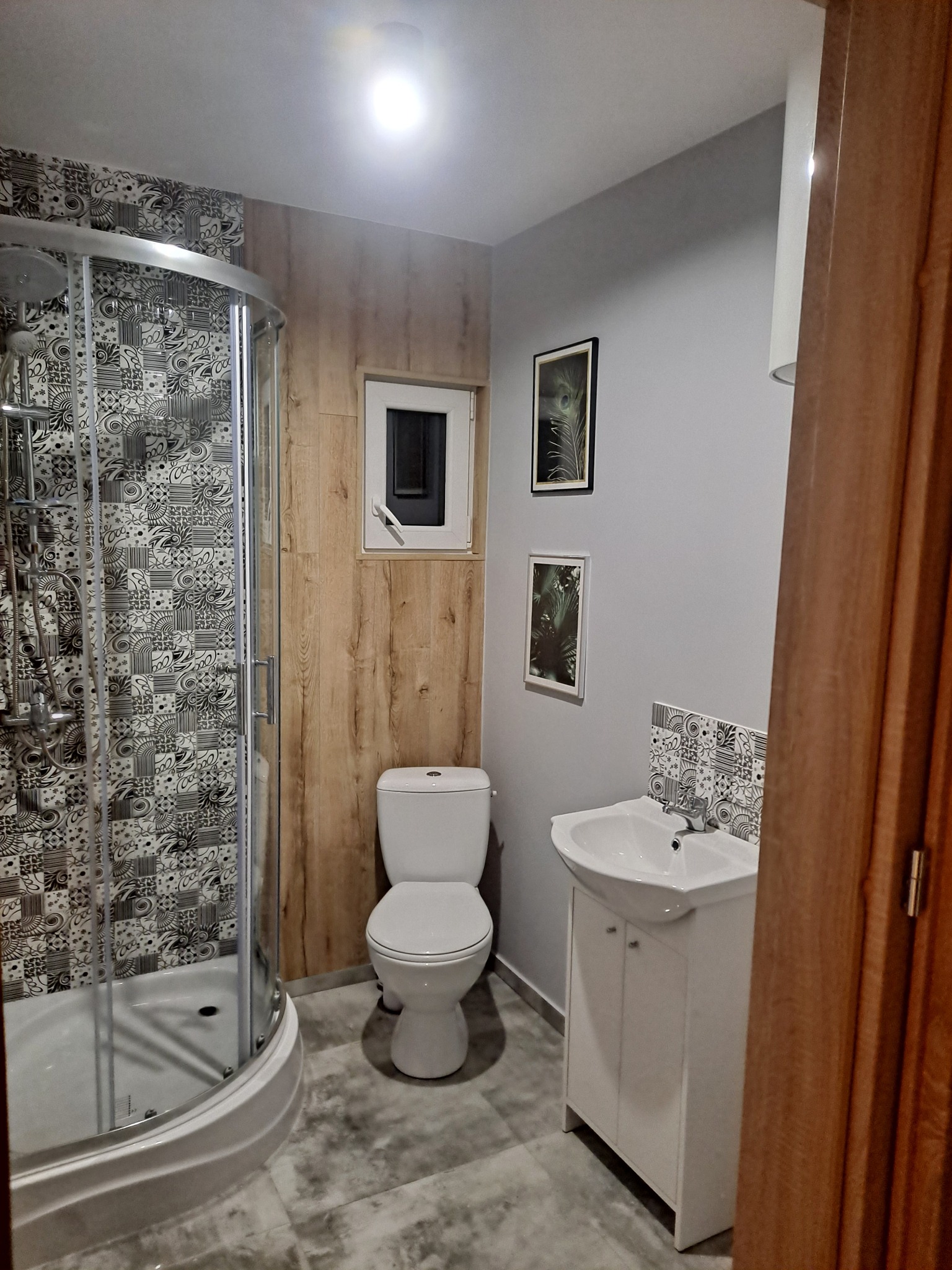 Wykończona łazienka w szarości i drewnie: kabina prysznicowa z wzorzystymi płytkami, toaleta, umywalka z szafką i drewniane panele ścienne.