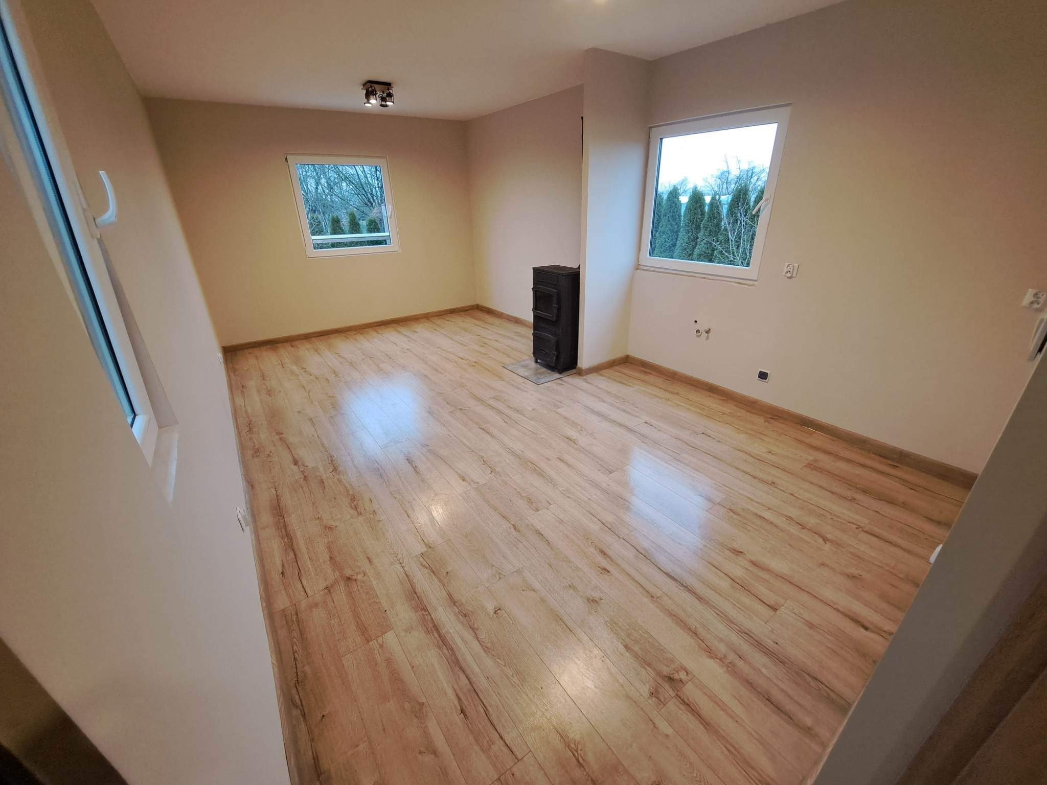 Pomalowane na beżowo puste pomieszczenie z panelami podłogowymi imitującymi drewno, czarnym piecem typu koza i dwoma oknami z widokiem na drzewa.