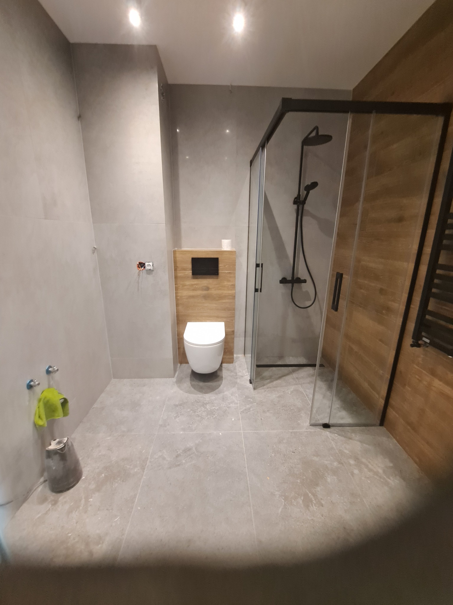 Nowoczesna łazienka z kabiną prysznicową typu walk-in z czarnymi ramami, podwieszaną toaletą i szarą płytką podłogową, widoczny fragment ściany z imitacją drewna oraz niebieskie uchwyty na ścianie...