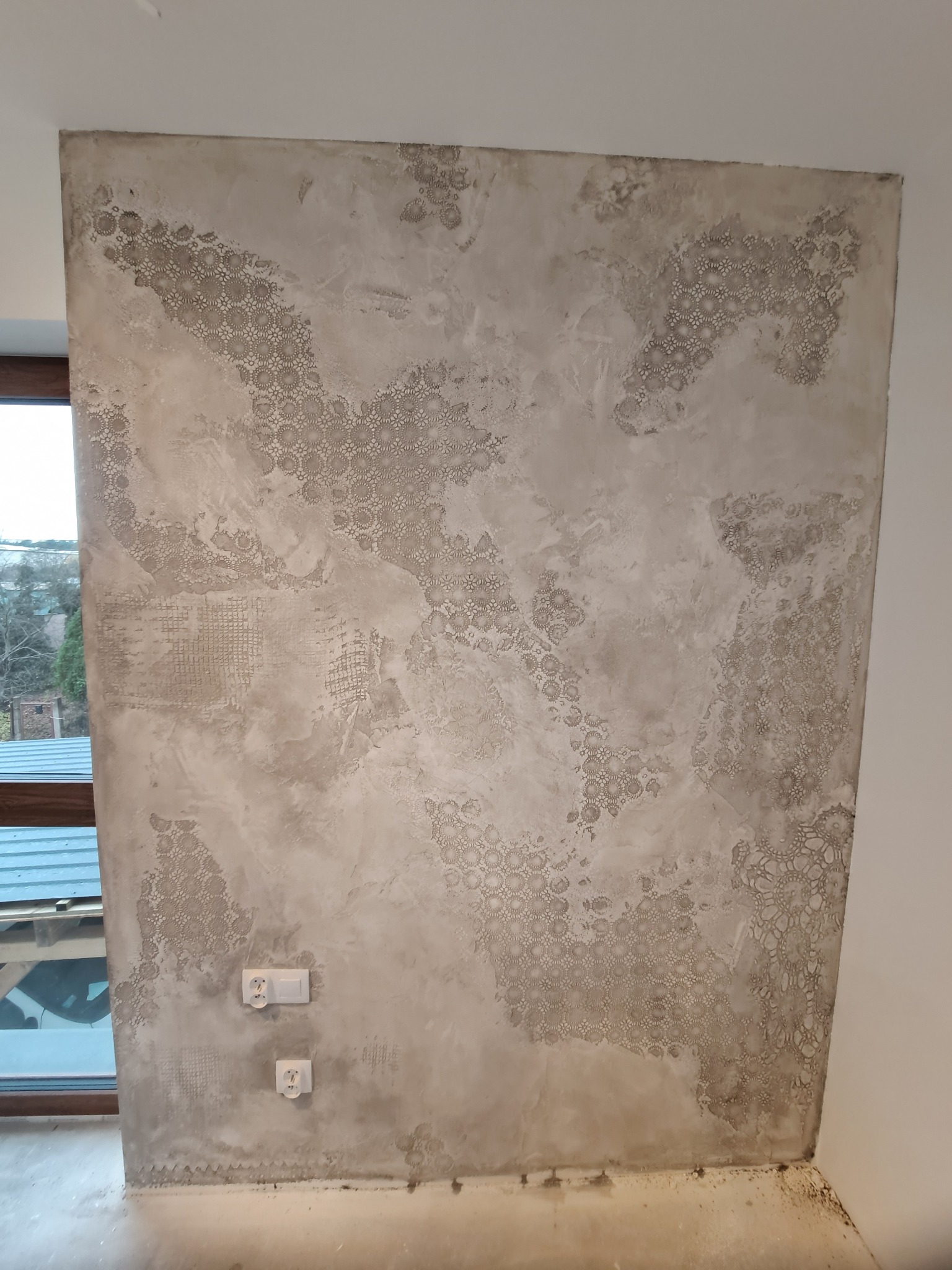 Ściana pokryta dekoracyjną, beżowo-szarą strukturą z motywem ażurowych rozet, widoczne gniazdka elektryczne, fragment okna z widokiem na zewnątrz.