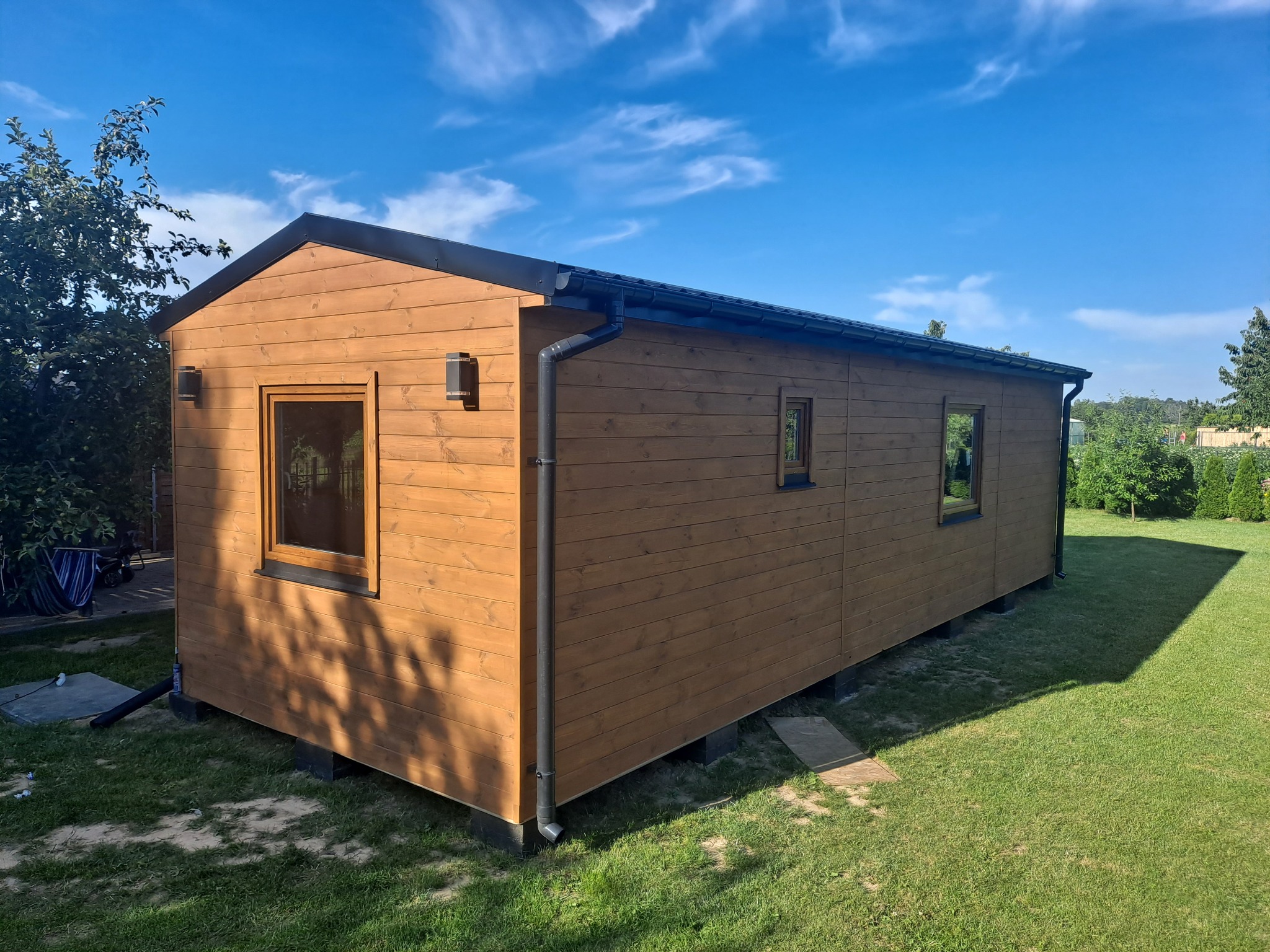 Nowoczesny, drewniany domek letniskowy z czarnym dachem i rynnami, osadzony na betonowych bloczkach, otoczony zielonym trawnikiem i drzewami w słoneczny dzień.