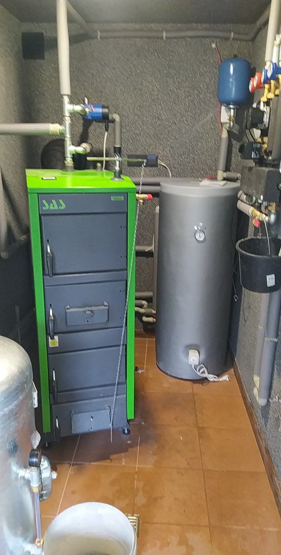 Kotłownia z piecem na paliwo stałe marki SAS w zielonej obudowie, zbiornikiem akumulacyjnym i instalacją hydrauliczną, widoczne ślady wody na podłodze z terakoty.