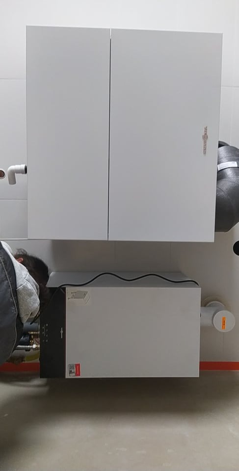 Instalacja białej jednostki wentylacyjnej z dwudrzwiową obudową, widoczny fragment instalatora przy podłączeniu przewodów, szare rury na ścianie z białych płytek.