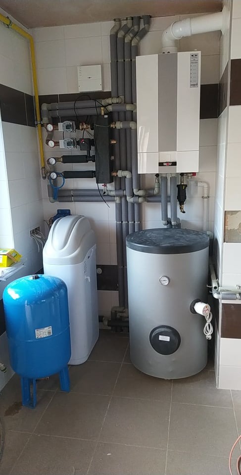 Zestaw urządzeń grzewczych w kotłowni: piec gazowy, zasobnik ciepłej wody, zmiękczacz wody i naczynie przeponowe, połączone rurami z izolacją termiczną.