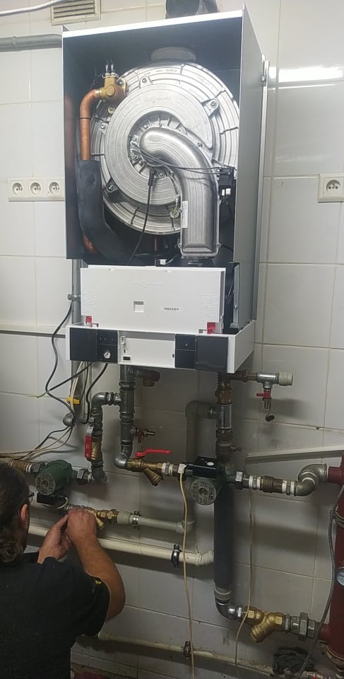 Instalator podłącza rury do nowoczesnego kotła grzewczego Viessmann na tle białych, kwadratowych płytek. Widoczne liczne zawory i elementy instalacji.