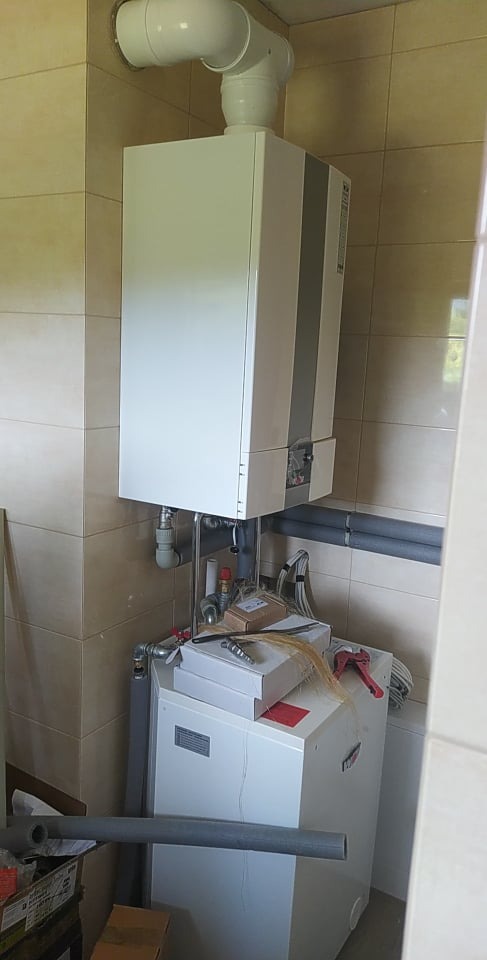 Biały piec grzewczy z podłączonymi rurami w pomieszczeniu wyłożonym beżowymi płytkami, na górze pieca leżą narzędzia i kartony.