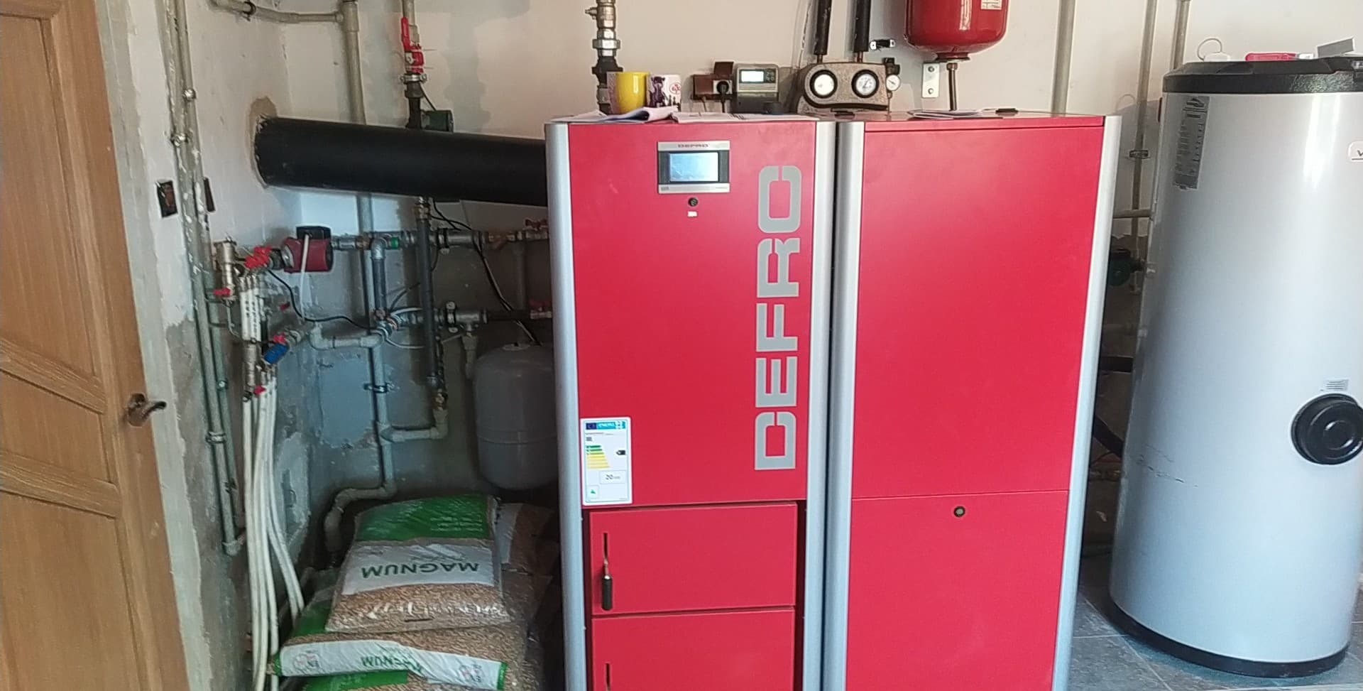 Czerwony piec na pellet marki DEFRO w kotłowni, obok zbiornik na wodę, rury instalacji grzewczej i zapas worków z pelletem Magnum.