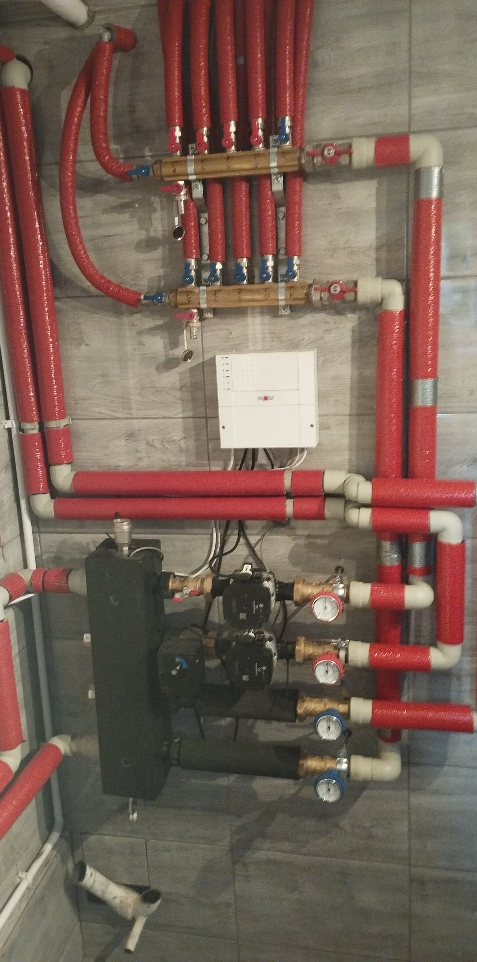 Rozdzielacz ogrzewania podłogowego z czerwonymi rurami izolowanymi, pompami obiegowymi i białą skrzynką sterującą na szarej ścianie z płytek.