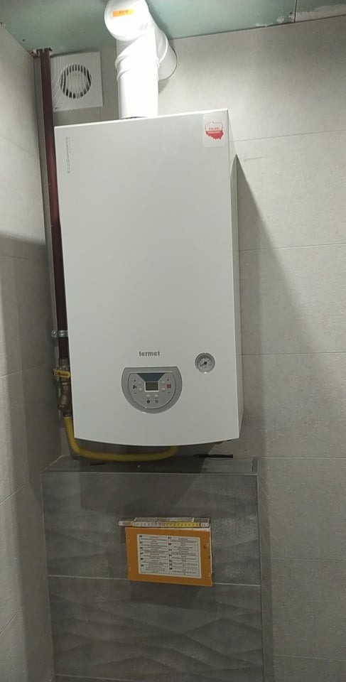 Biały piec gazowy marki Termet, zamontowany na szarej, kafelkowanej ścianie, z widoczną instalacją gazową i kominem odprowadzającym spaliny, dodatkowo widoczna kratka wentylacyjna i dokumentacja...