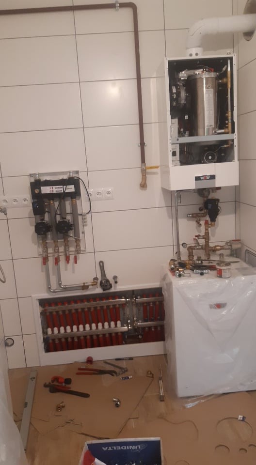 Wnętrze kotłowni z zamontowanym kotłem grzewczym, rozdzielaczem podłogówki i osprzętem hydraulicznym na tle białych płytek. Na podłodze widoczne narzędzia i karton.