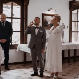 Elegancko ubrana para, kobieta w białej sukni ciążowej, mężczyzna w szarym garniturze z muszką, wznoszą toast w przestronnym wnętrzu z dużymi oknami i drewnianymi ramami, w tle stoi mężczyzna...