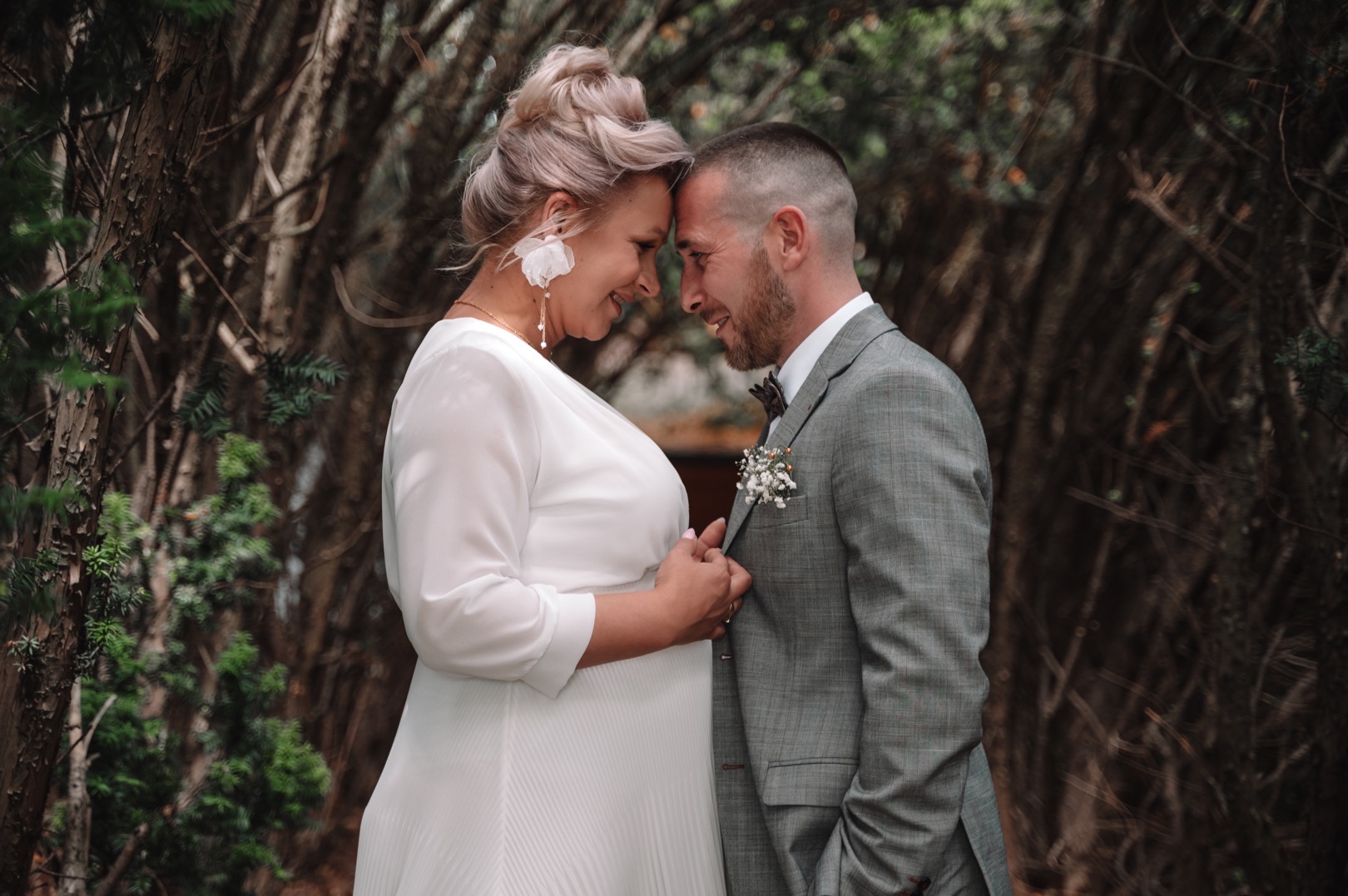 Para młoda, kobieta w białej sukni z plisowanym dołem i kwiatowych kolczykach, mężczyzna w szarym garniturze w kratę z butonierką, stykają się czołami na tle gęstych krzewów.