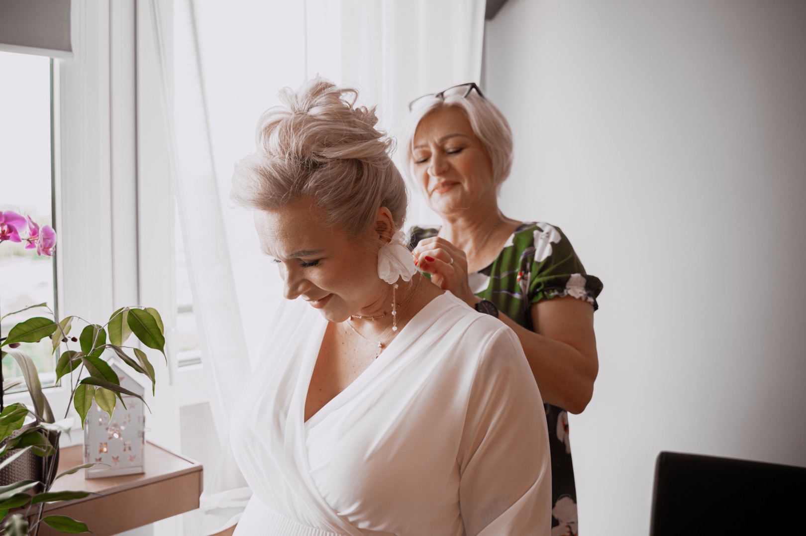 Kobieta w białej sukience z dekoltem w serek, poprawiana przez drugą kobietę w kwiecistej bluzce, w tle okno z storczykiem i dekoracyjna latarenka.