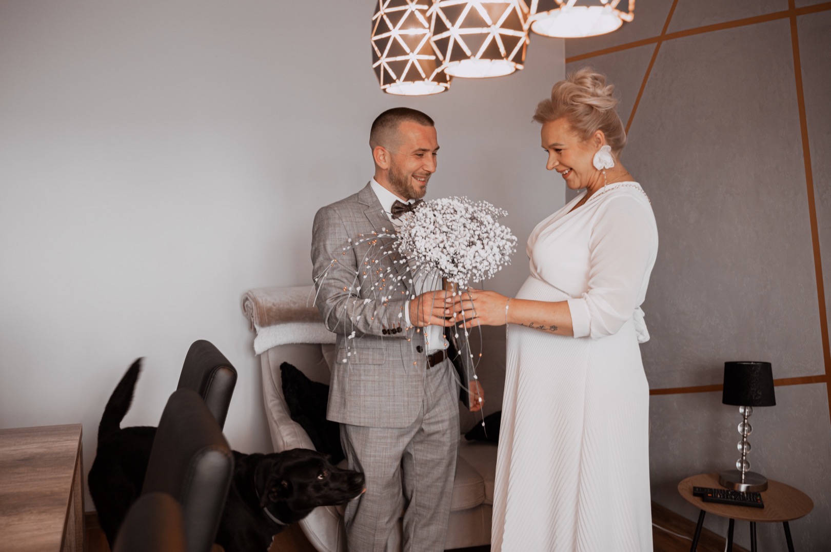 Uśmiechnięty mężczyzna w szarym garniturze z muszką wręcza bukiet gipsówki ciężarnej kobiecie w białej sukience, czarny pies patrzy z dołu, nowoczesne lampy wiszą nad nimi.