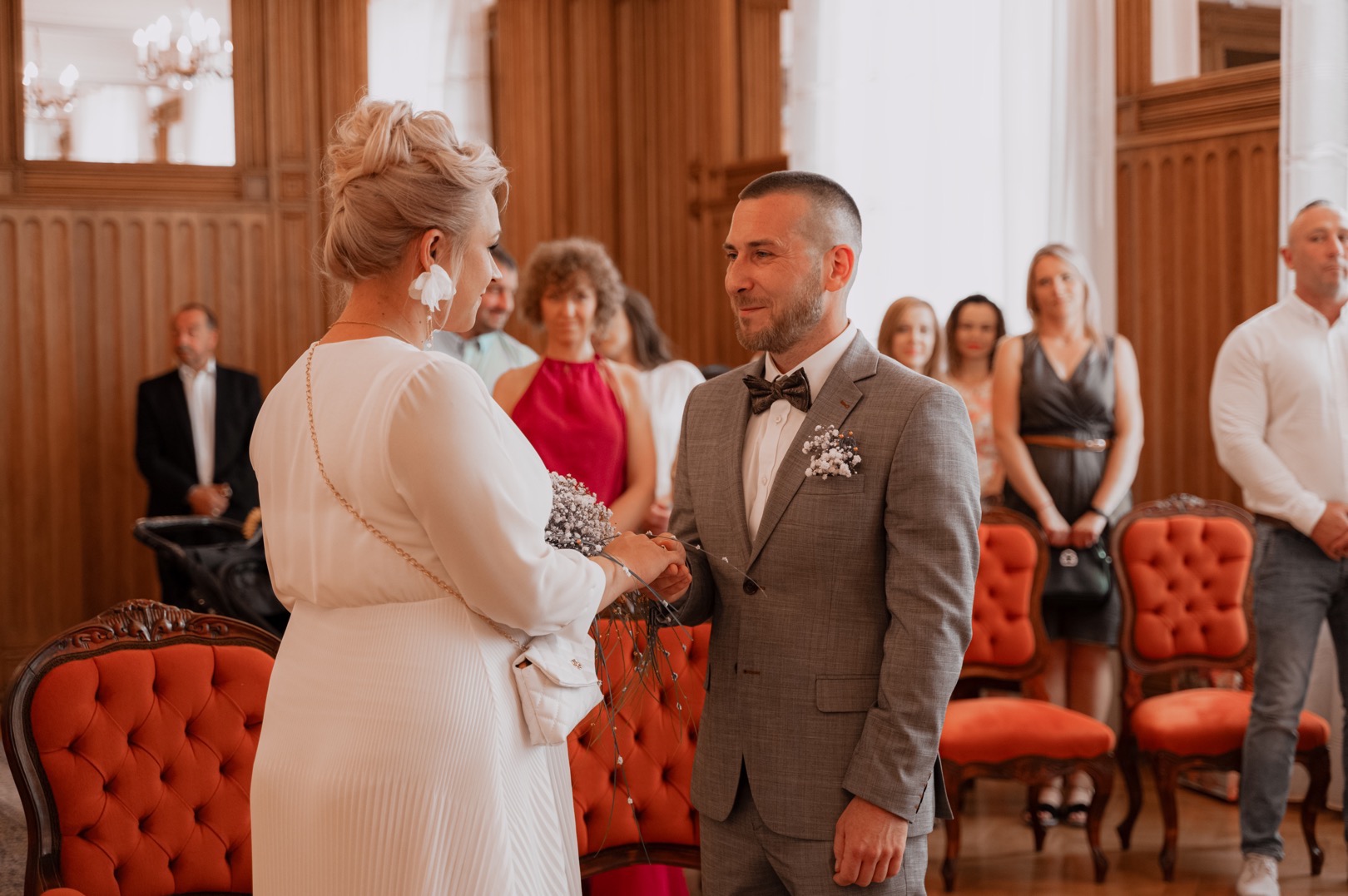 Para młoda wymienia się obrączkami podczas ceremonii ślubnej w eleganckim wnętrzu, z gośćmi w tle i dekoracyjnymi krzesłami.