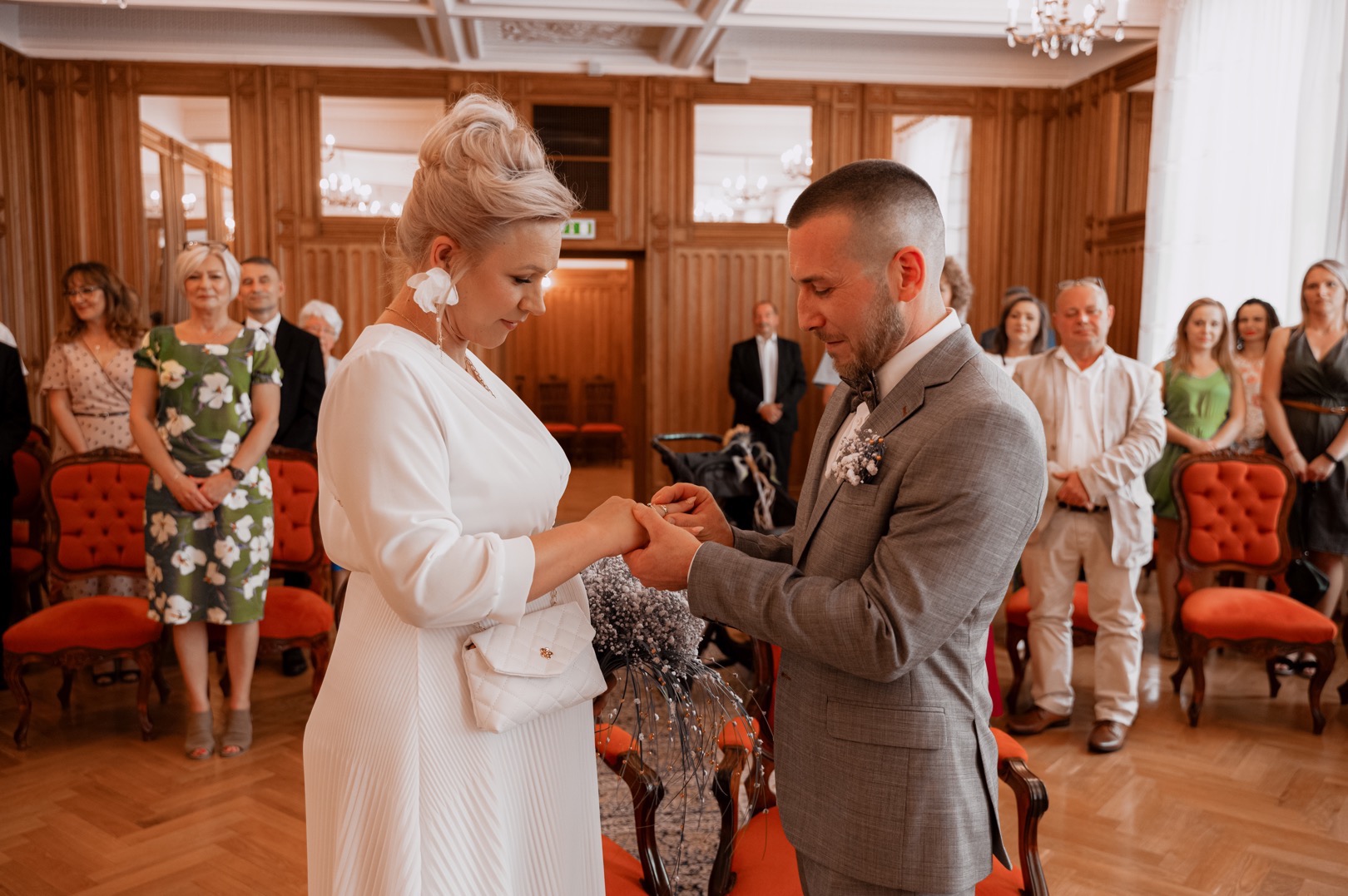 Para wymienia się obrączkami podczas ceremonii ślubnej w eleganckim wnętrzu z drewnianymi panelami i żyrandolami, na tle obserwujących gości.