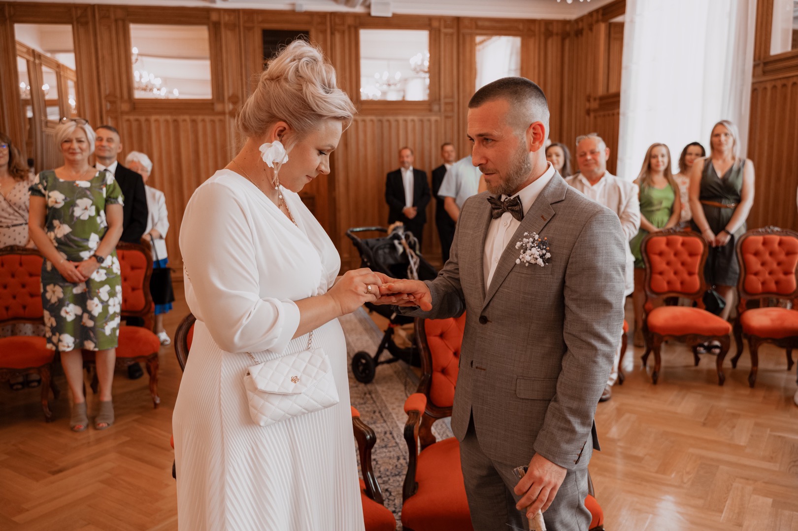 Panna młoda w białej sukni zakłada obrączkę na palec pana młodego w szarym garniturze podczas ceremonii ślubnej w eleganckim wnętrzu, w tle goście i krzesła z czerwonym obiciem.