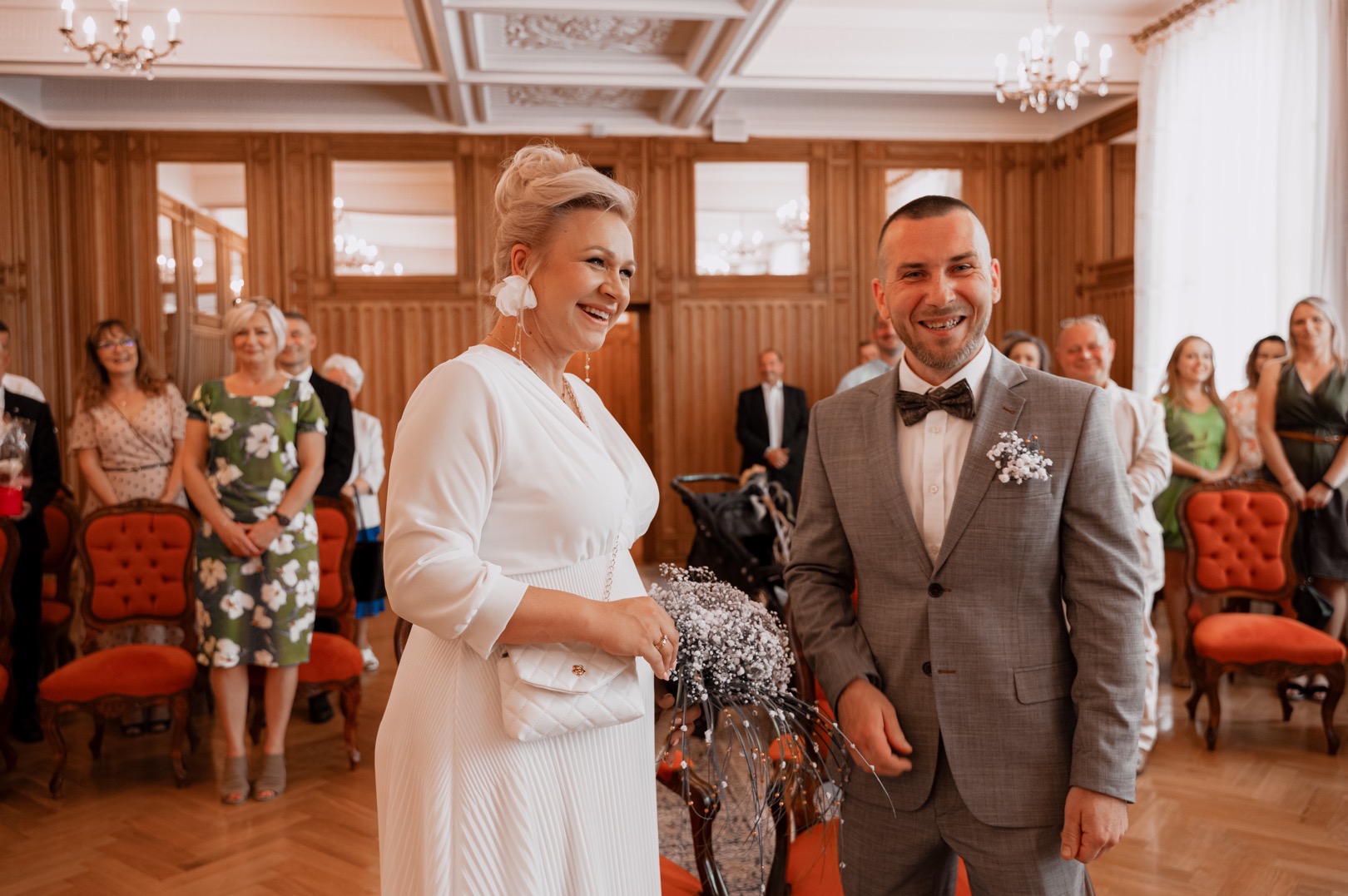 Uśmiechnięta para młoda w eleganckich strojach podczas ceremonii ślubnej, ona w białej sukience z plisowanym dołem i torebką Chanel, on w szarym garniturze i muszce, goście obserwujący w tle.