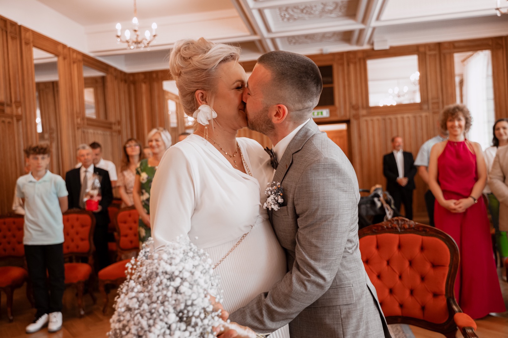 Pocałunek przyszłych rodziców w eleganckim wnętrzu, ona w białej sukni ciążowej z bukietem gipsówki, on w szarym garniturze, w tle goście.