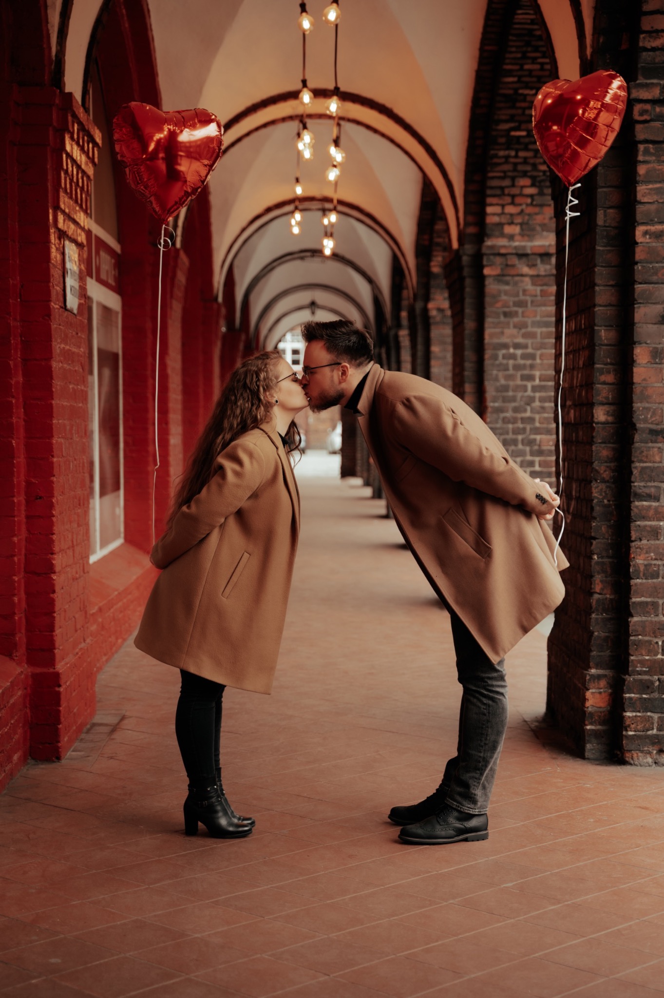 Para całująca się w arkadach, z czerwonymi balonami w kształcie serc, oświetlona wiszącymi żarówkami, oboje w beżowych płaszczach.
