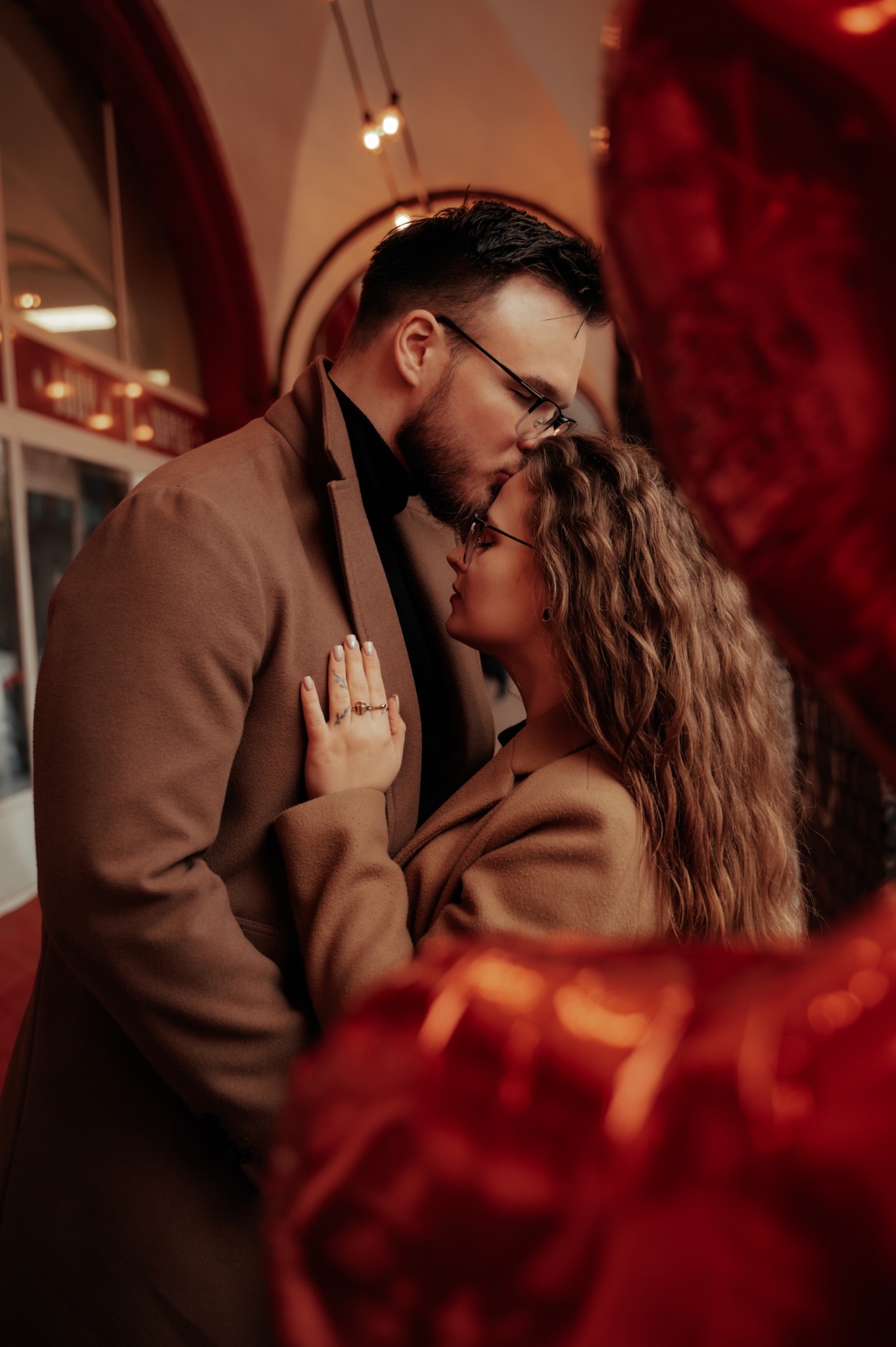 Mężczyzna w okularach czule całuje kobietę w czoło, oboje w brązowych płaszczach, w tle czerwone balony i oświetlona witryna.