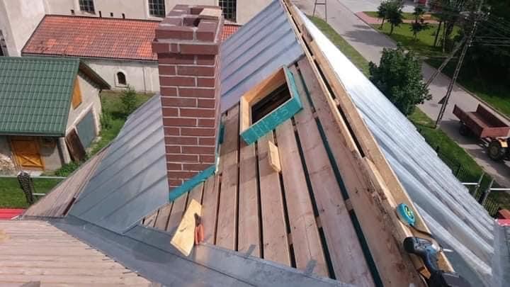 Montaż dachu z blachy trapezowej, widoczne drewniane łaty, okno dachowe w trakcie montażu i komin z cegły, w tle zabudowa wiejska.