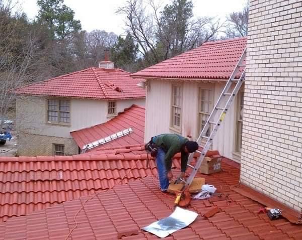 Dekarz w zielonej koszuli i niebieskich jeansach naprawia czerwoną dachówkę na dachu budynku z cegły, drabina oparta o ścianę, w tle budynki z czerwonymi dachami i drzewa.