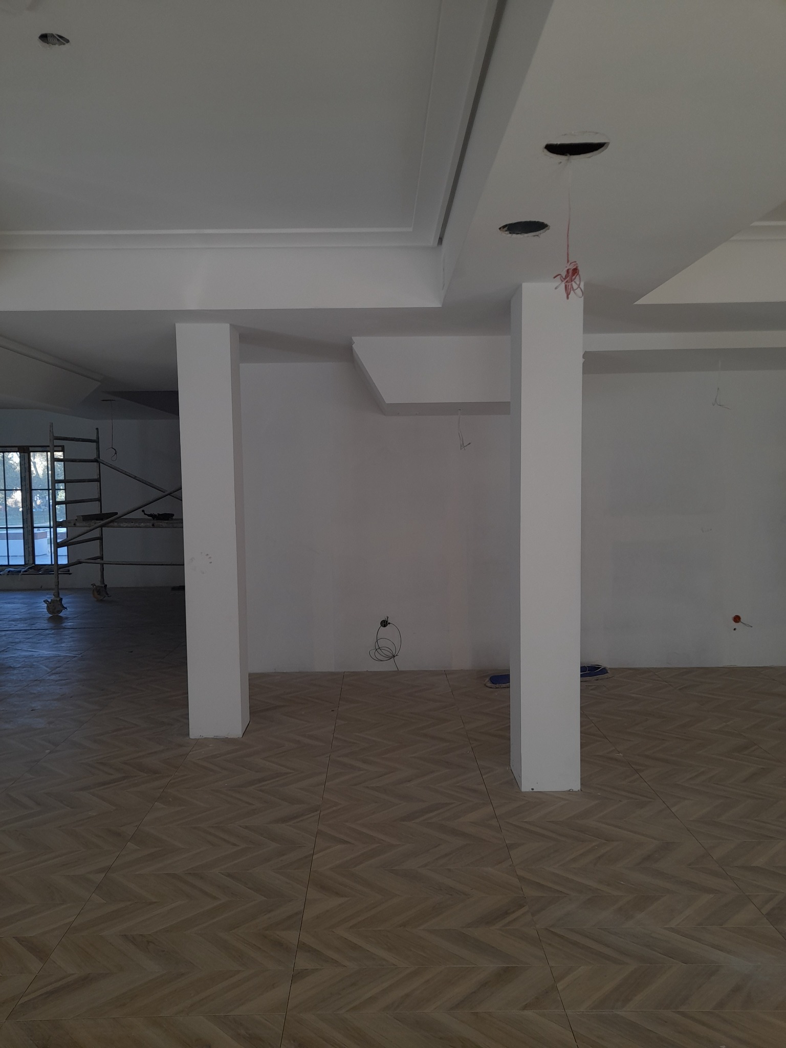Wnętrze w trakcie prac wykończeniowych: widoczne kolumny, ułożona podłoga w jodełkę, sufit z otworami na oświetlenie i wystające kable, ściany przygotowane pod malowanie, rusztowanie przy oknie.