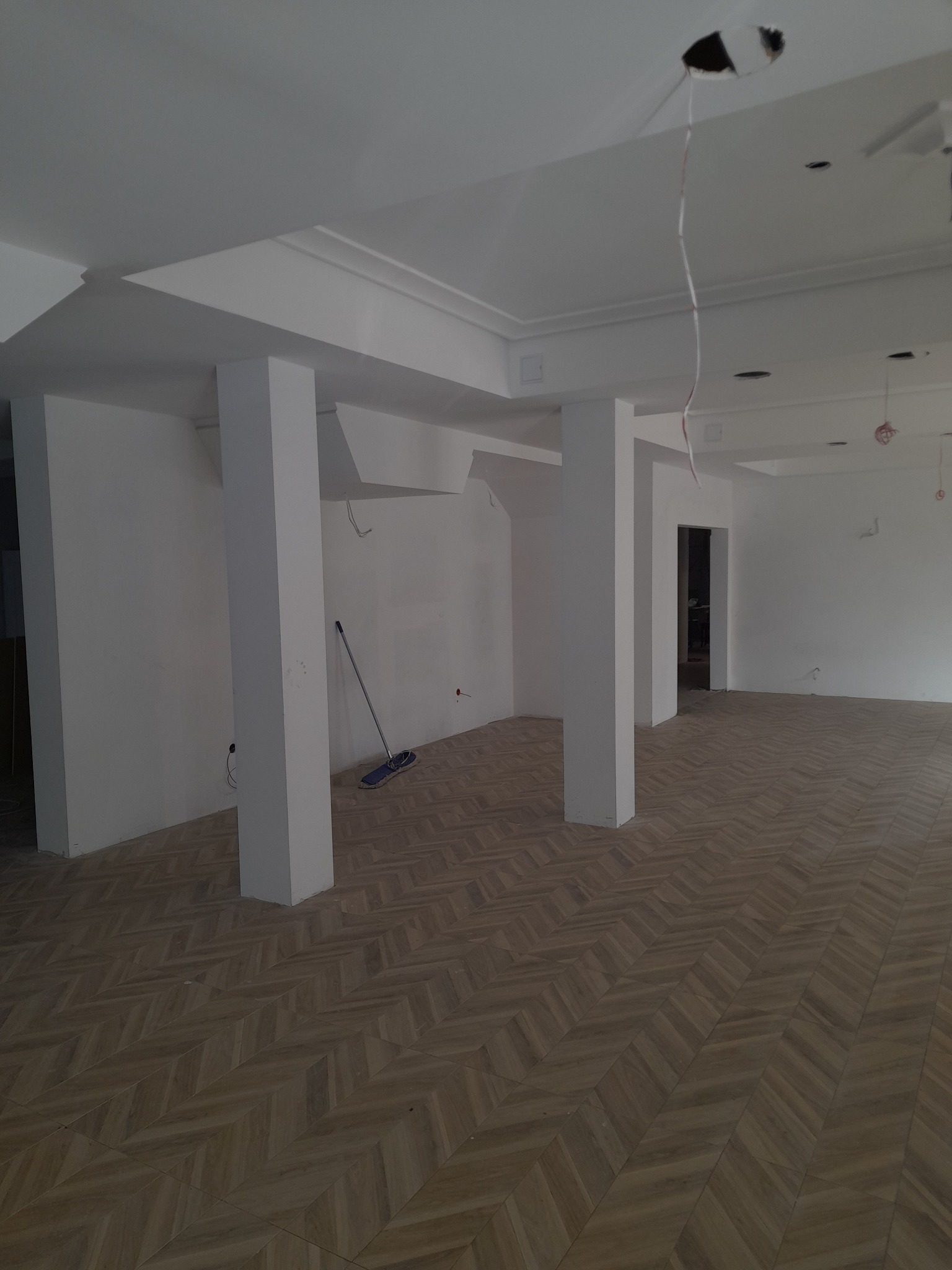 Wnętrze biura w trakcie wykańczania: białe ściany, kolumny, parkiet w jodełkę, widoczne niedokończone instalacje elektryczne i mop oparty o ścianę.