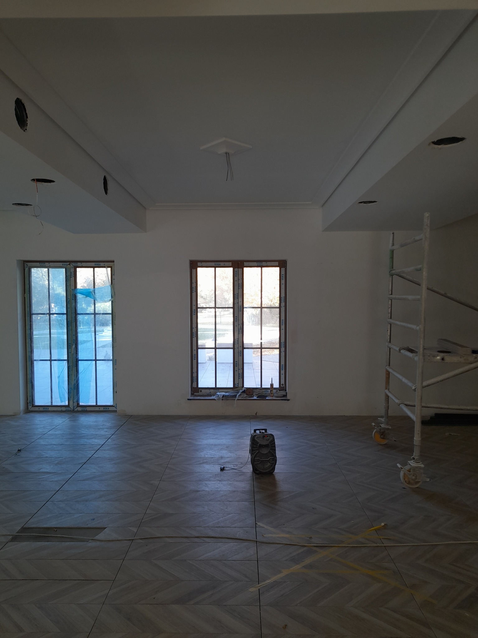Wnętrze w trakcie remontu: ułożona podłoga z płytek w jodełkę, pomalowane na biało ściany z oknami zabezpieczonymi folią, widoczne otwory na oświetlenie w suficie oraz rusztowanie malarskie...