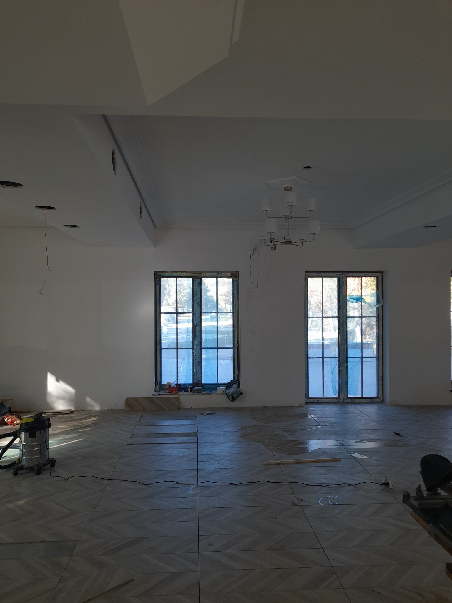 Układanie parkietu w jodełkę w przestronnym, jasnym pokoju z dwoma dużymi oknami zabezpieczonymi folią, widoczny odkurzacz przemysłowy i narzędzia glazurnicze.