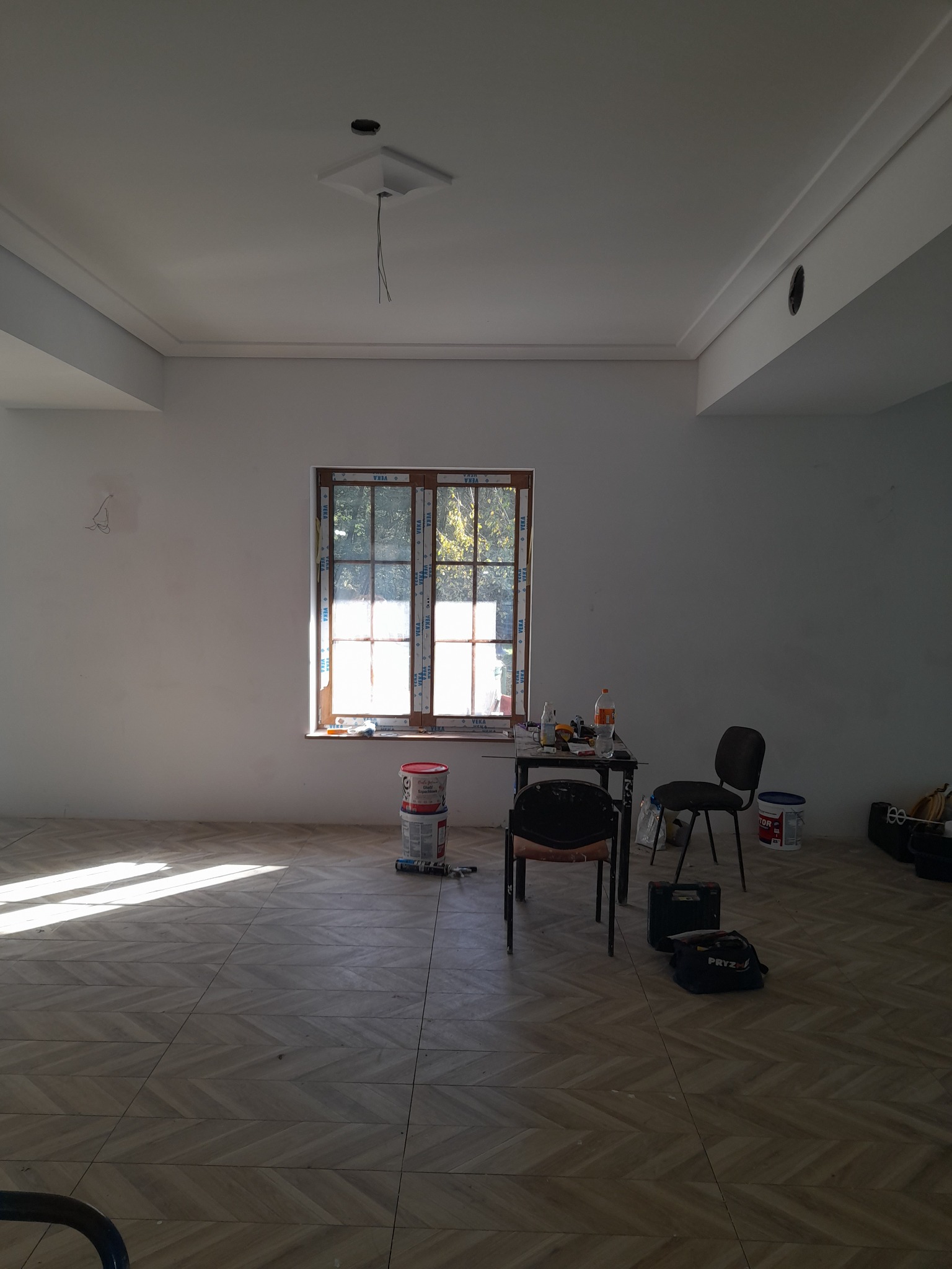 Wnętrze pokoju w trakcie prac wykończeniowych, widoczne puszki z farbą, narzędzia, krzesła i okno z widokiem na zewnątrz. Na podłodze ułożona jest jasna, wzorzysta terakota.