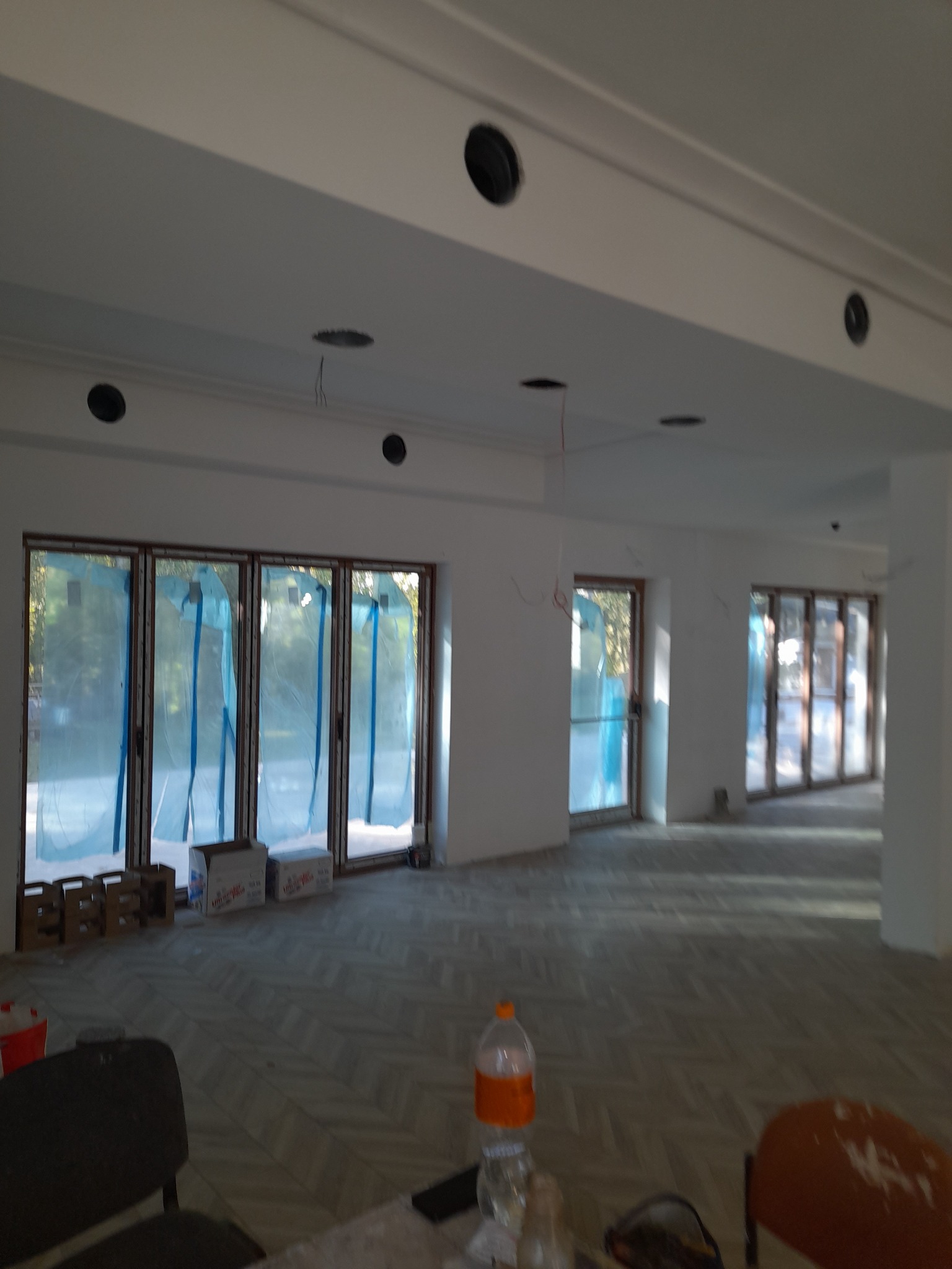 Wnętrze w trakcie prac wykończeniowych: widoczne okna zabezpieczone folią, otwory w suficie pod oświetlenie, ułożona podłoga i puszki po materiałach budowlanych.