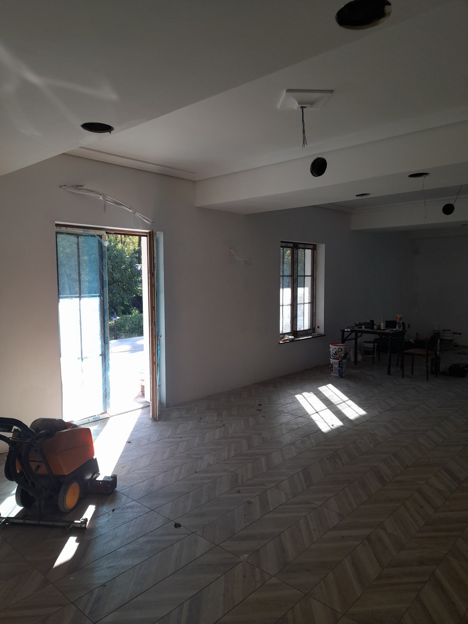 Wnętrze w trakcie remontu z nową podłogą w jodełkę, świeżo pomalowanymi ścianami, otwartymi drzwiami balkonowymi i widocznymi otworami w suficie na oświetlenie, maszyna do czyszczenia podłóg stoi...