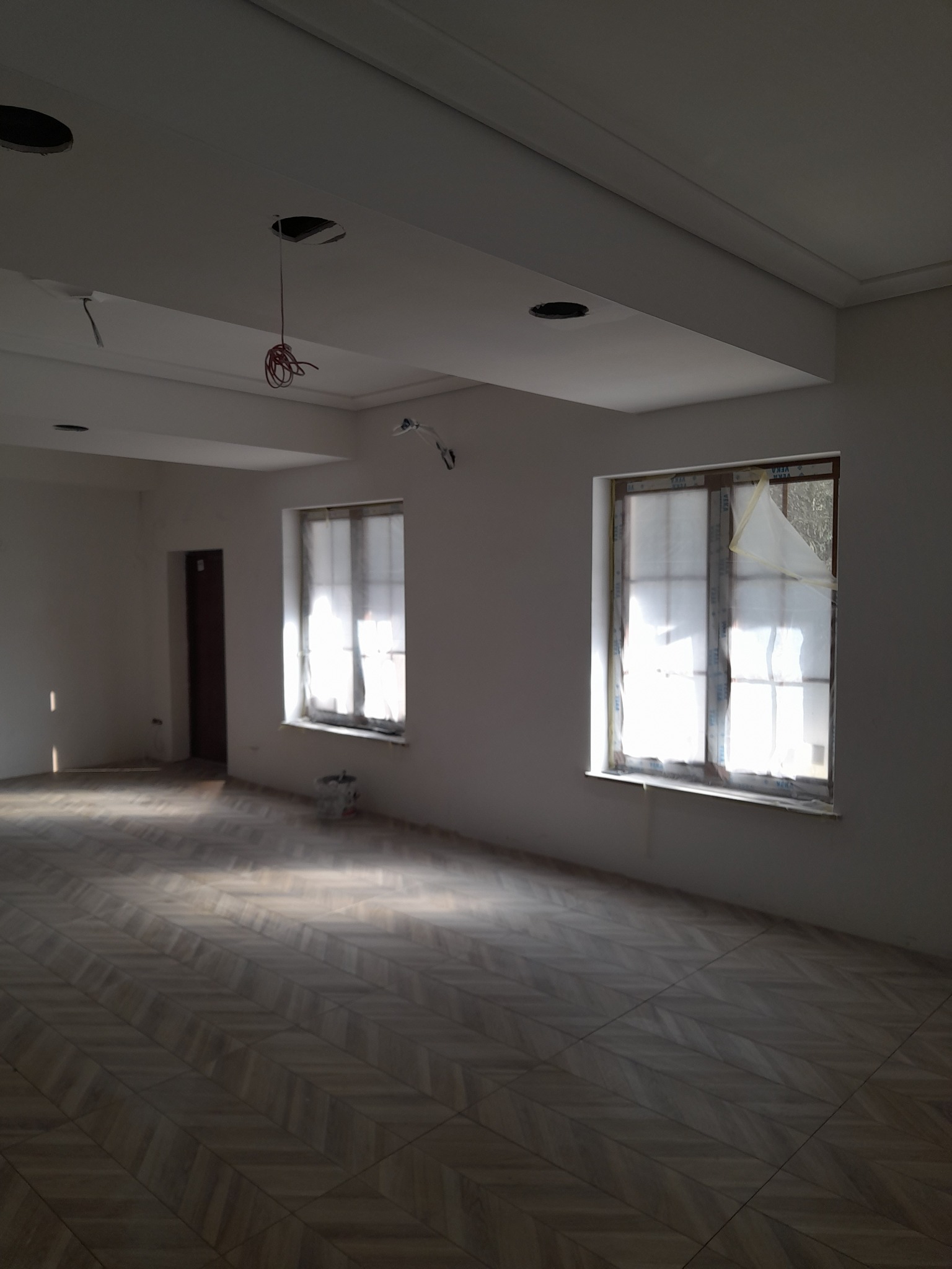 Wnętrze w trakcie prac wykończeniowych: świeżo pomalowane ściany w jasnym odcieniu, okna zabezpieczone folią malarską, parkiet ułożony we wzór jodełki, widoczne przewody elektryczne wystające...