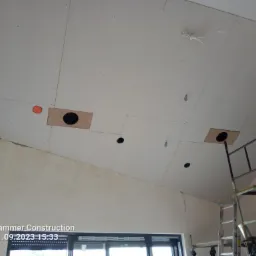 Skośny sufit z płyt kartonowo-gipsowych z widocznymi otworami na oświetlenie i wentylację, częściowo wykończony, z widocznym rusztowaniem i elementami instalacji elektrycznej.