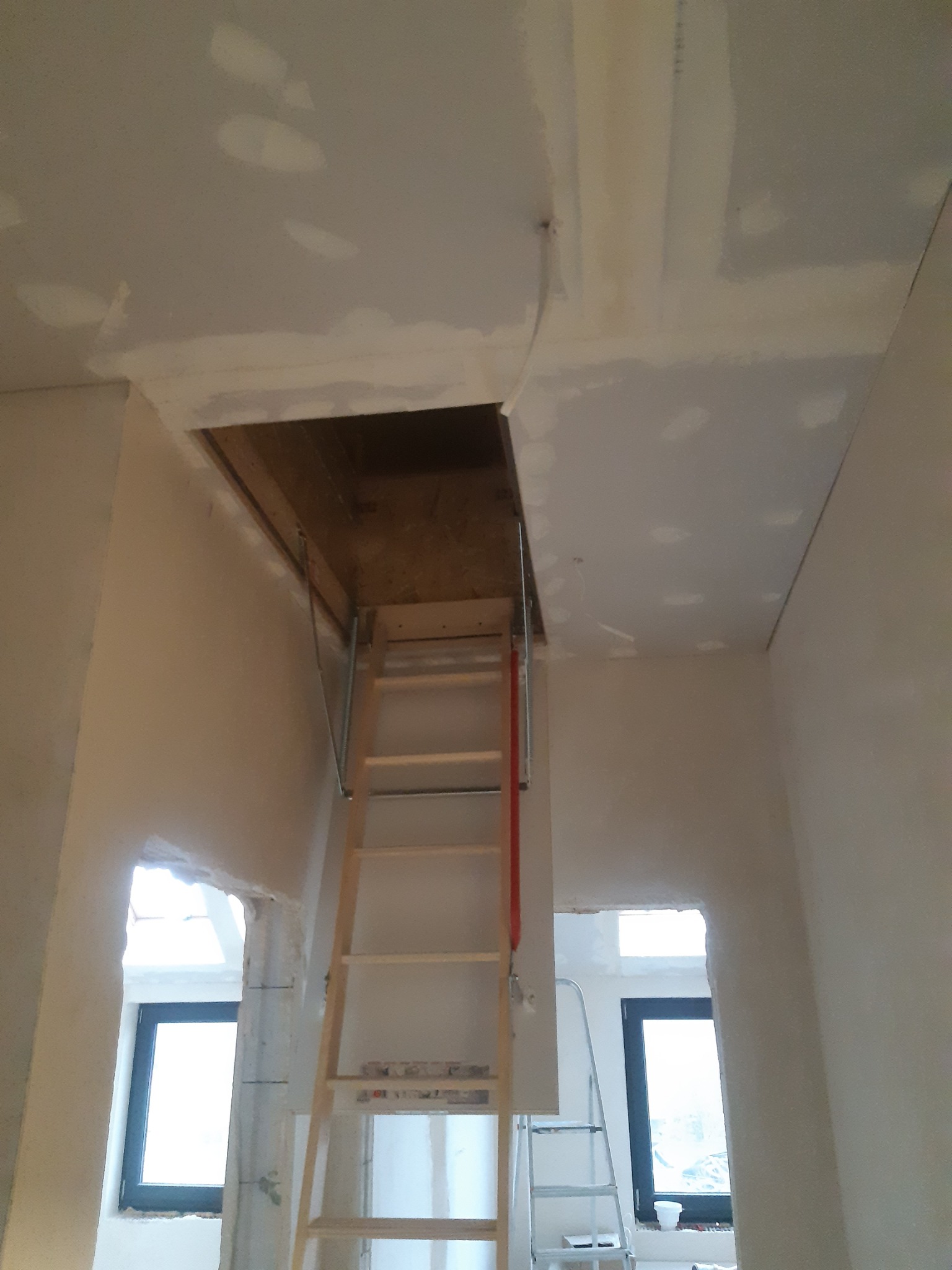 Montaż składanych schodów strychowych w suficie z widocznymi łączeniami płyt gipsowo-kartonowych przed szpachlowaniem, w tle okno i fragment drabiny.