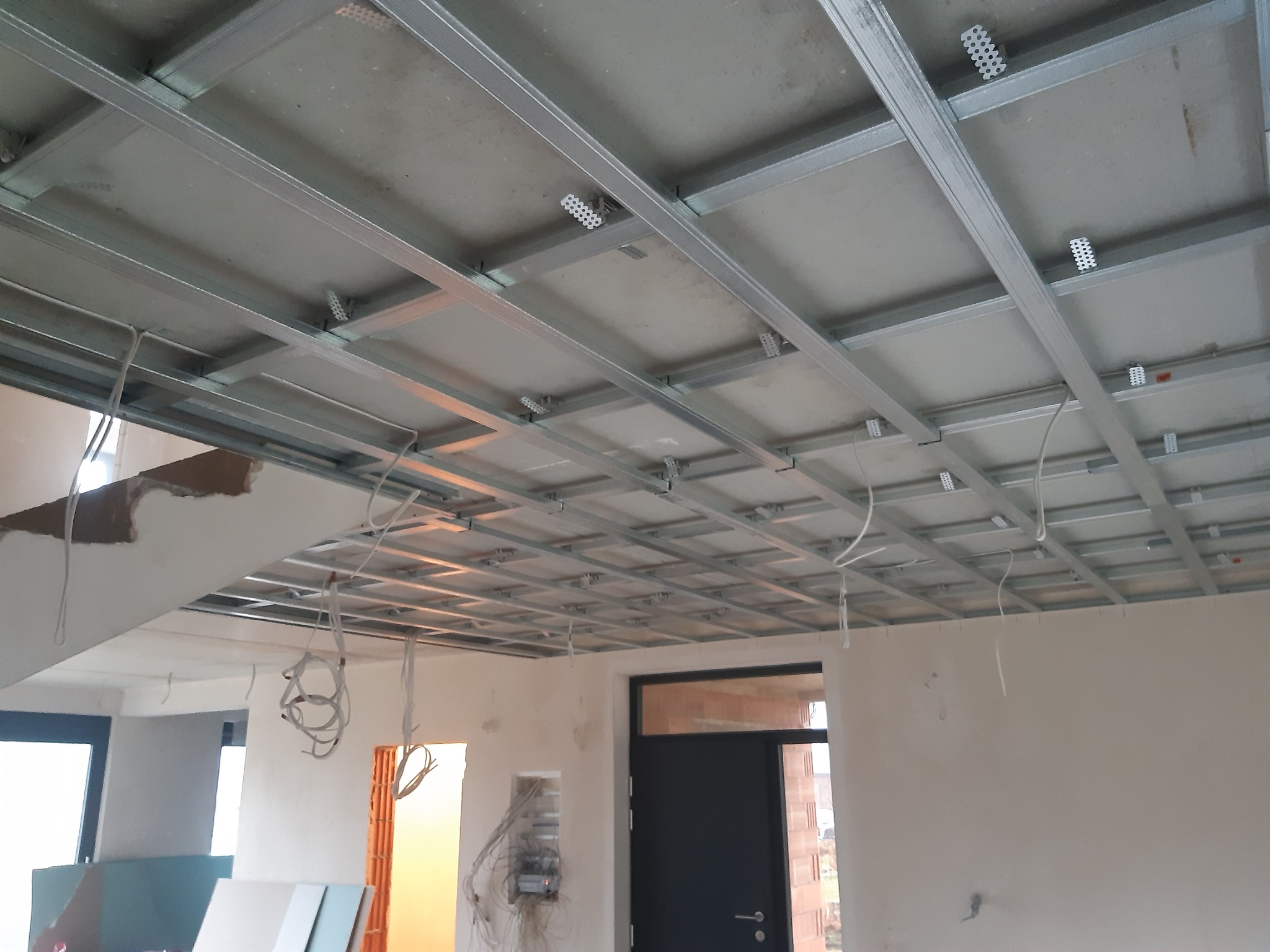 Stelaż sufitu podwieszanego z profili metalowych, częściowo obłożony płytami, widoczne wiszące przewody elektryczne i niedokończone ściany w surowym stanie.