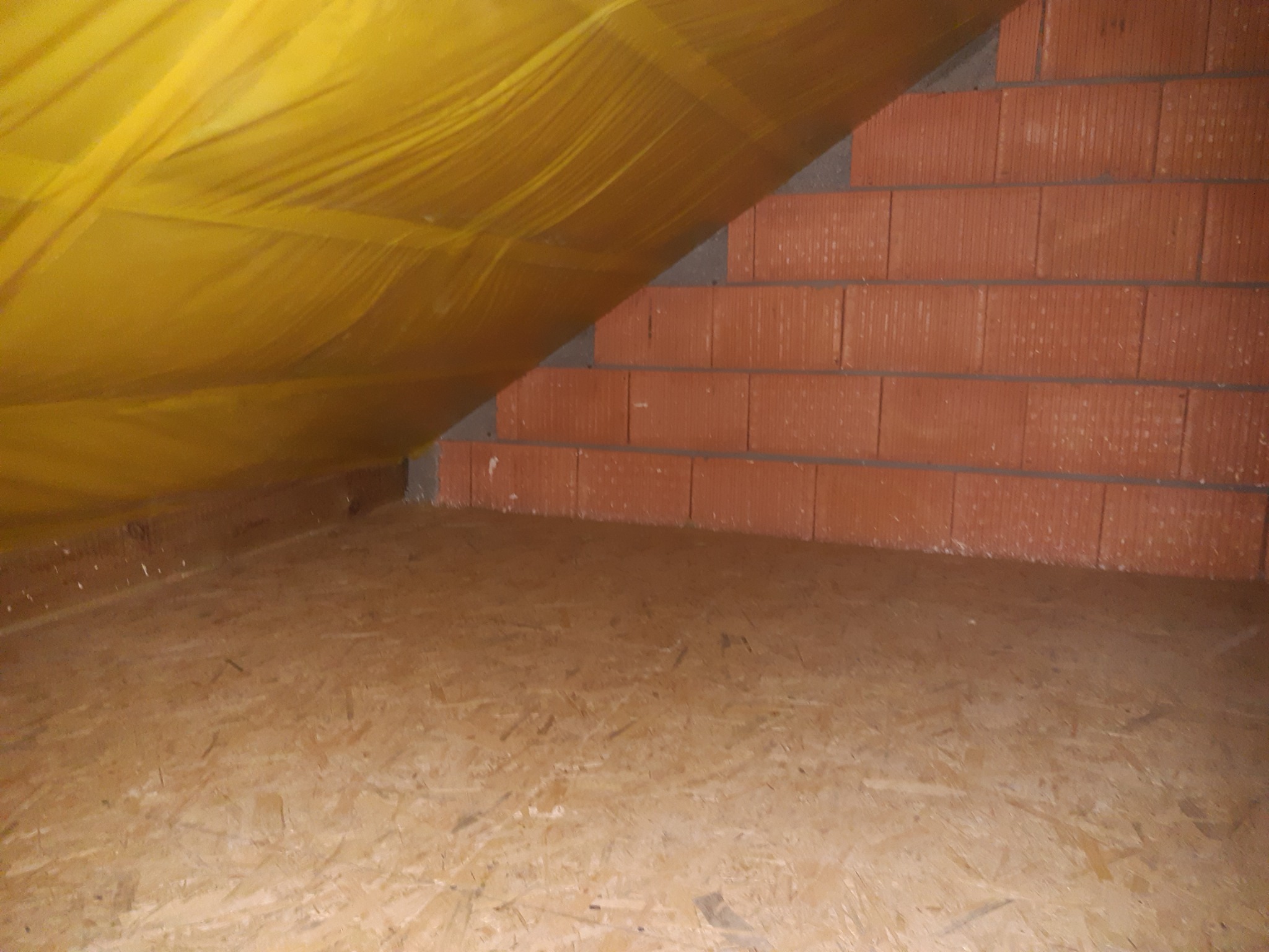 Wnętrze poddasza w trakcie prac: widoczna żółta folia izolacyjna, fragment ściany z czerwonej cegły oraz podłoga z płyty OSB.