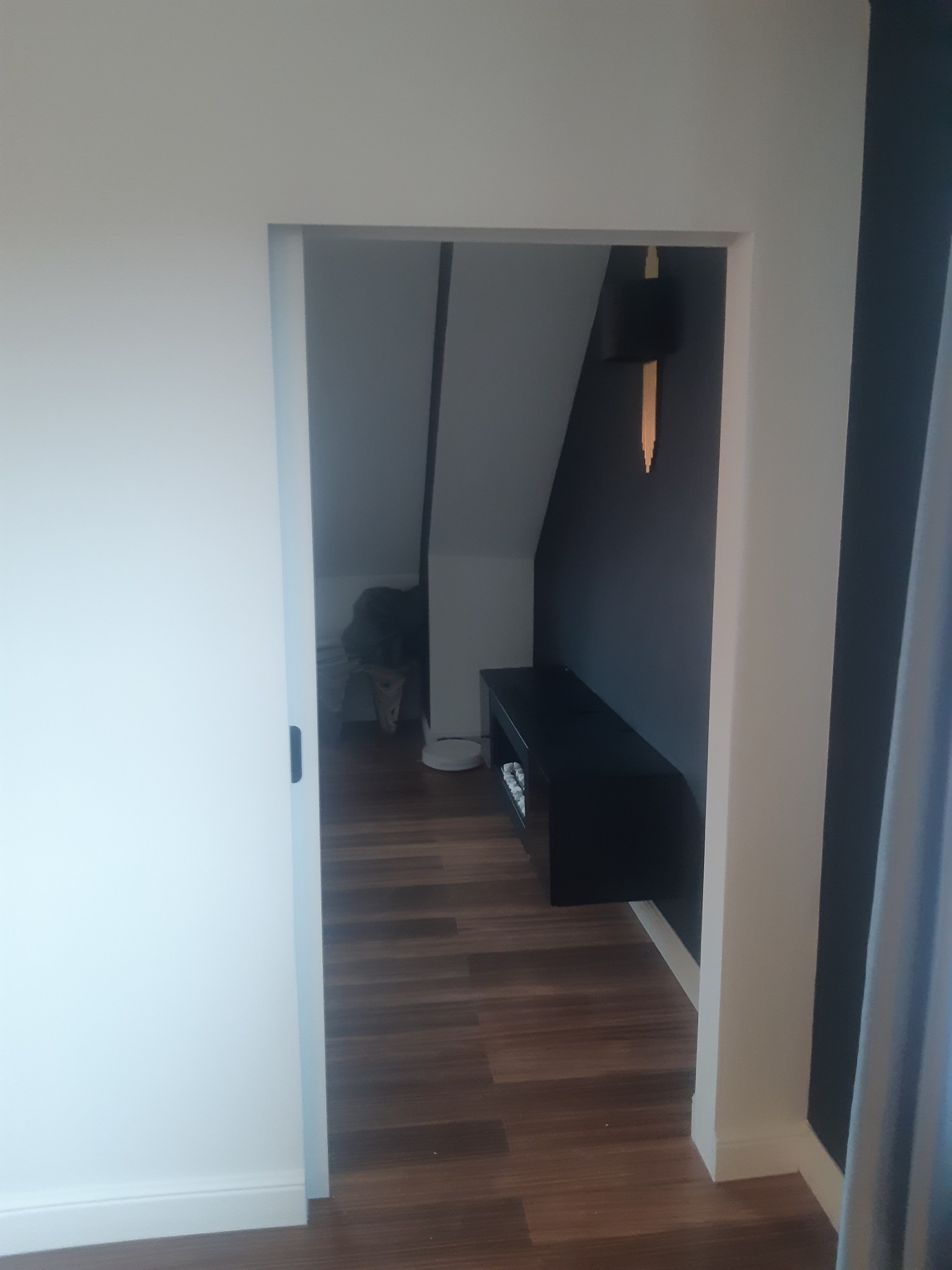 Widok na wykończone poddasze przez otwarte drzwi przesuwne, z ciemną ścianą, półką i ozdobną lampą, oraz widocznym odkurzaczem automatycznym na podłodze.