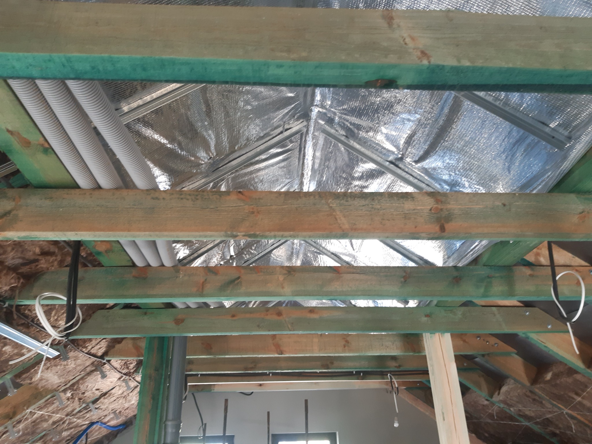 Srebrna folia izolacyjna na poddaszu, widoczna spod konstrukcji drewnianej z zielonymi elementami, z poprowadzonymi rurami instalacyjnymi