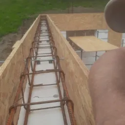 Zbrojenie w szalunku fundamentowym z płyt OSB, widoczne elementy izolacji termicznej i wystające pręty zbrojeniowe w tle.